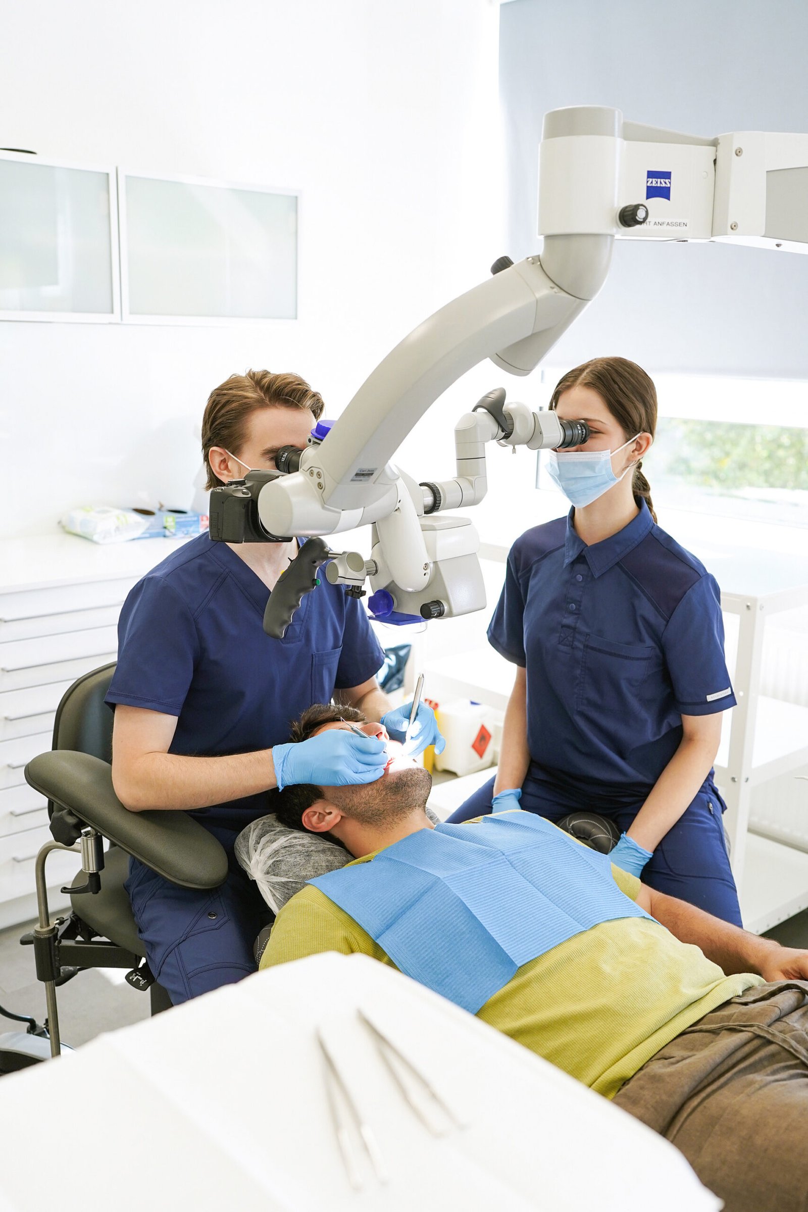 Zahnarzt behandelt einen Patienten unter dem ZEISS-Operationsmikroskop, assistiert von einer Mitarbeiterin